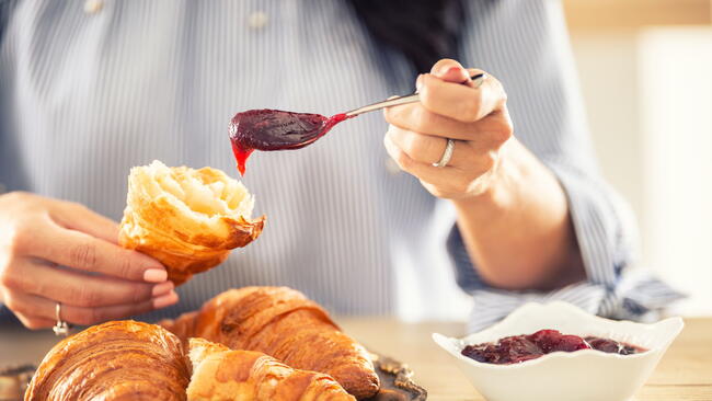 Female hands put jam onto a halved croissant in the morning.